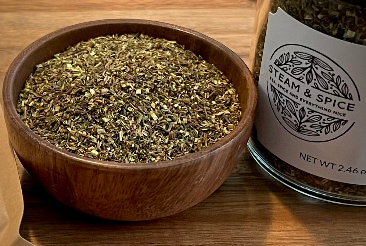 A wooden bowl filled with loose Green Rooibos next to a glass jar labeled Steam & Spice tea, on a wooden tabletop.
