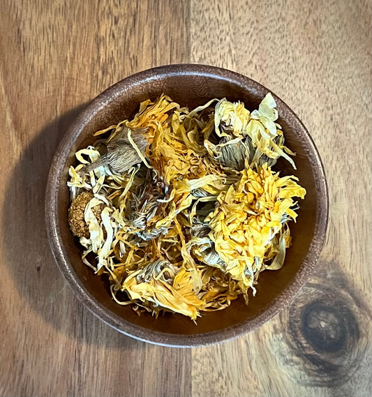 Dried yellow and white flowers, likely chamomile or calendula, piled in a small brown ceramic bowl on a wooden tabletop.