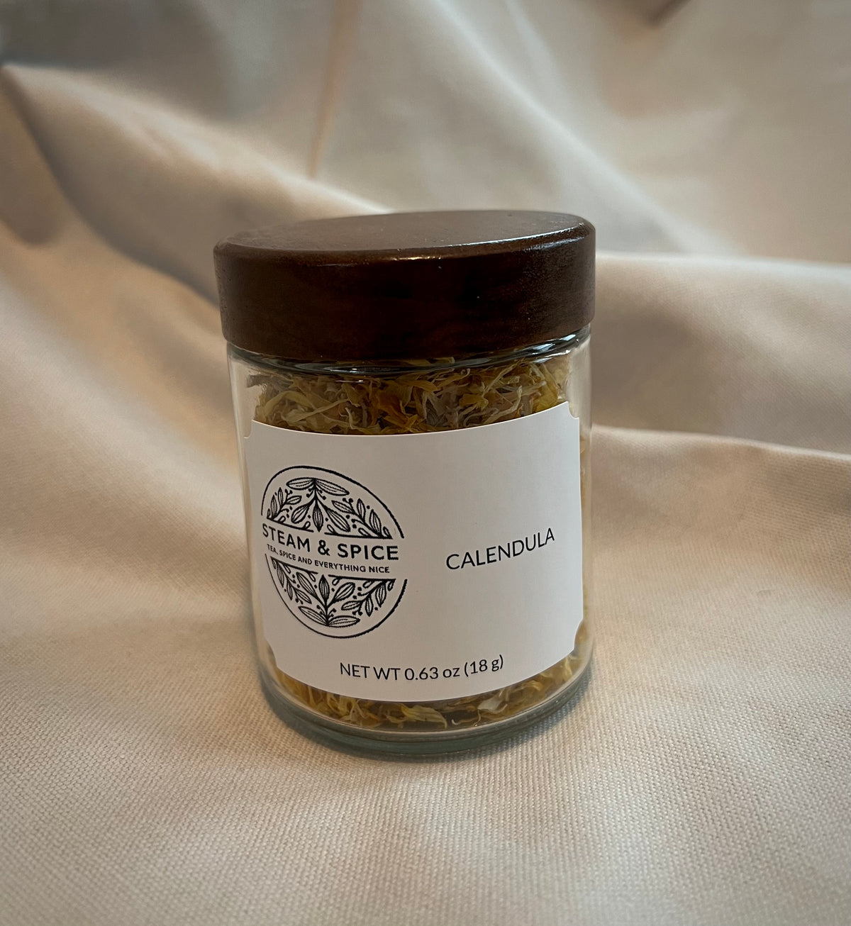 A clear glass jar of Calendula herbal tea with a dark brown wooden lid and a white label, set against a draped beige fabric background.