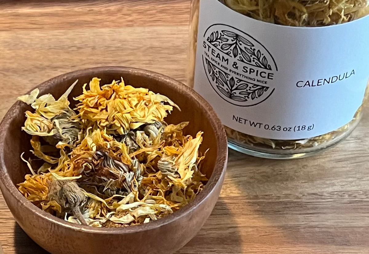 A wooden bowl filled with dried calendula flowers sits in front of a glass jar labeled 'Steam & Spice' and 'Calendula' on a wooden tabletop.