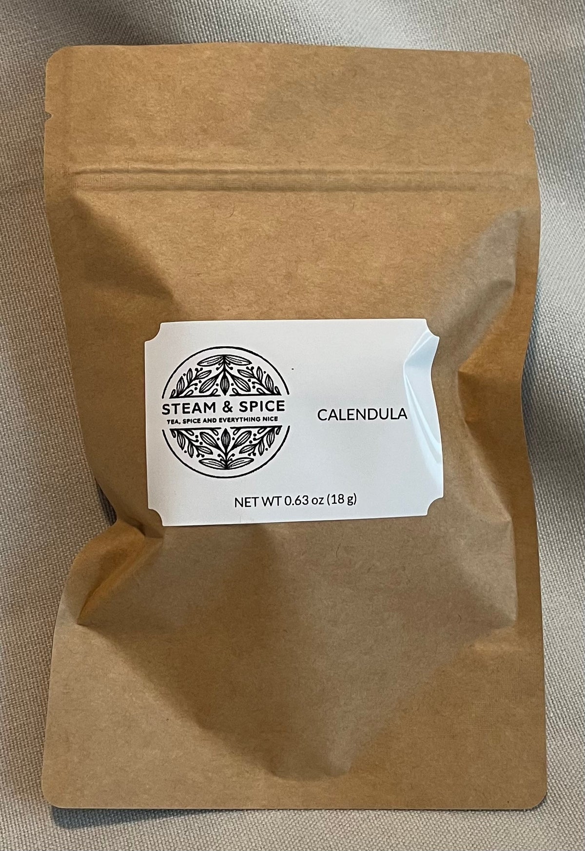A brown kraft paper stand-up pouch with a white label reading 'STEAM & SPICE' and 'CALENDULA' on a neutral fabric background.