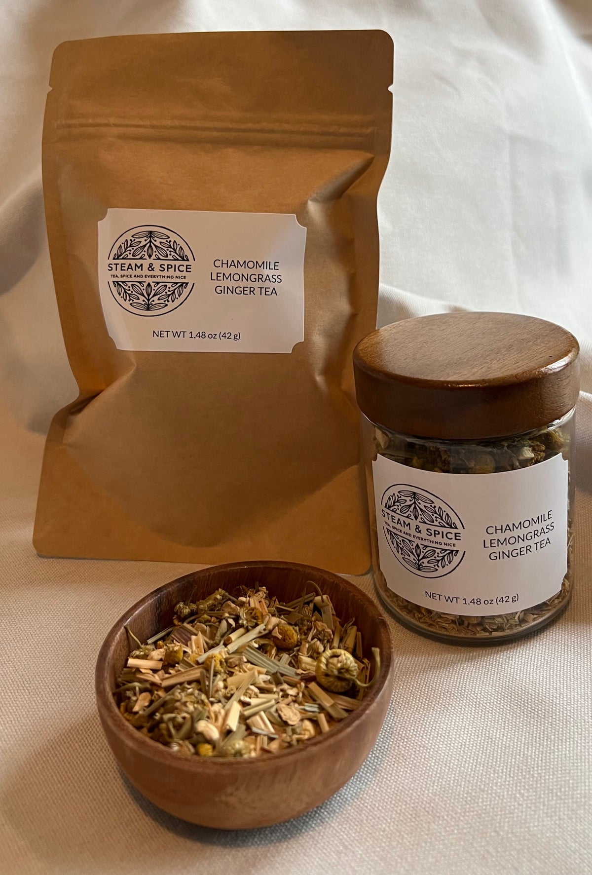 A kraft paper pouch and a glass jar of Steam & Spice Chamomile Lemongrass Ginger Tea are displayed with a small wooden bowl of loose tea leaves in front, set against a neutral draped fabric background.