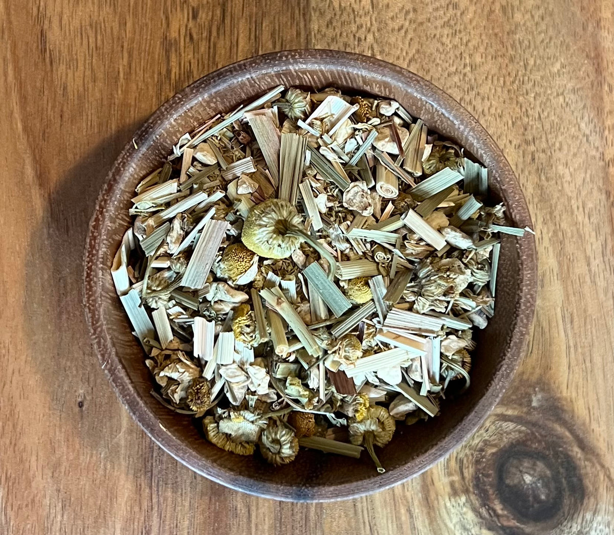 A top-down view of Chamomile, Lemongrass Ginger herbal tea blend, including chamomile flowers and leafy stems, presented in a round brown wooden bowl on a wooden tabletop.