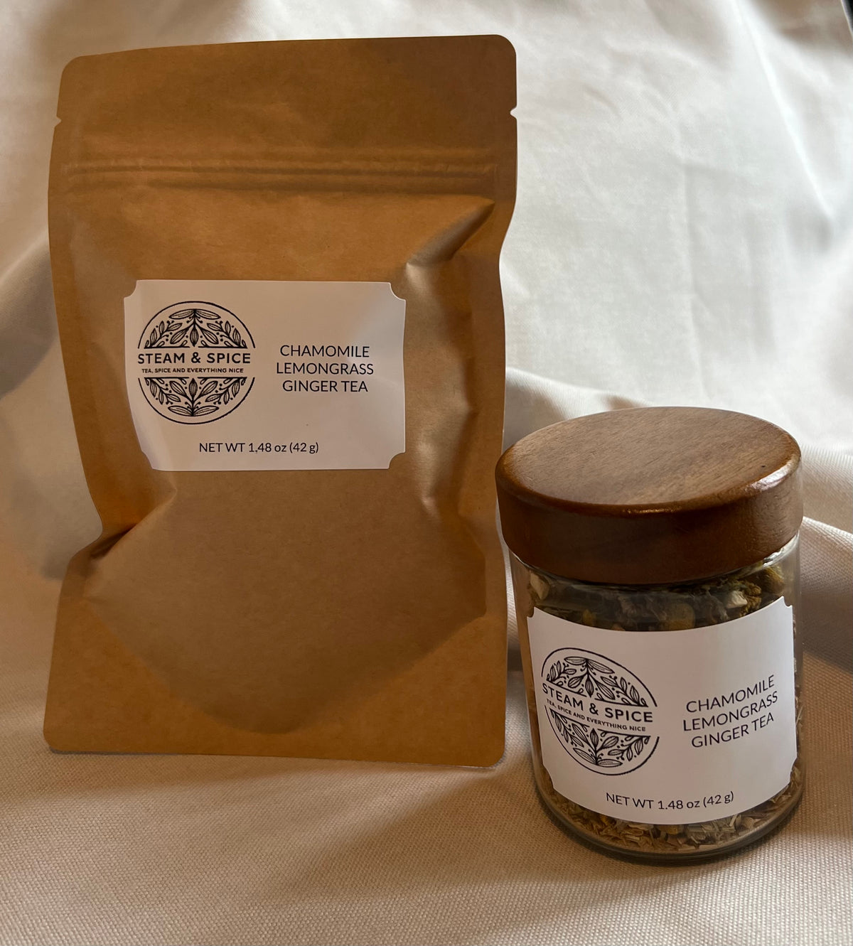A kraft paper stand-up pouch and a small glass jar of chamomile lemongrass ginger tea from Steam & Spice, both with white labels, displayed on a draped cream fabric background.