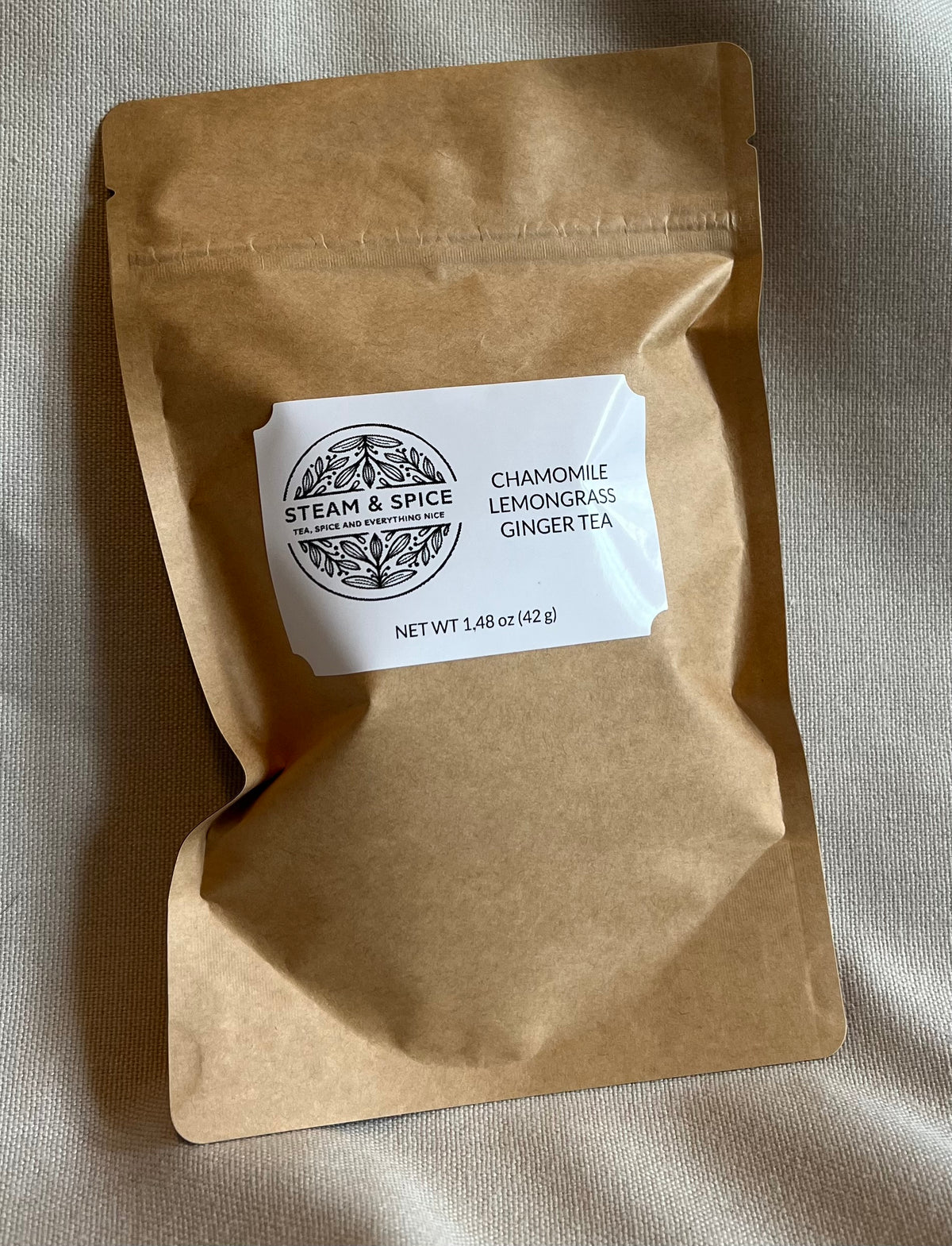 A kraft paper stand-up pouch of Steam & Spice Chamomile Lemongrass Ginger Tea, placed on a textured beige fabric background.