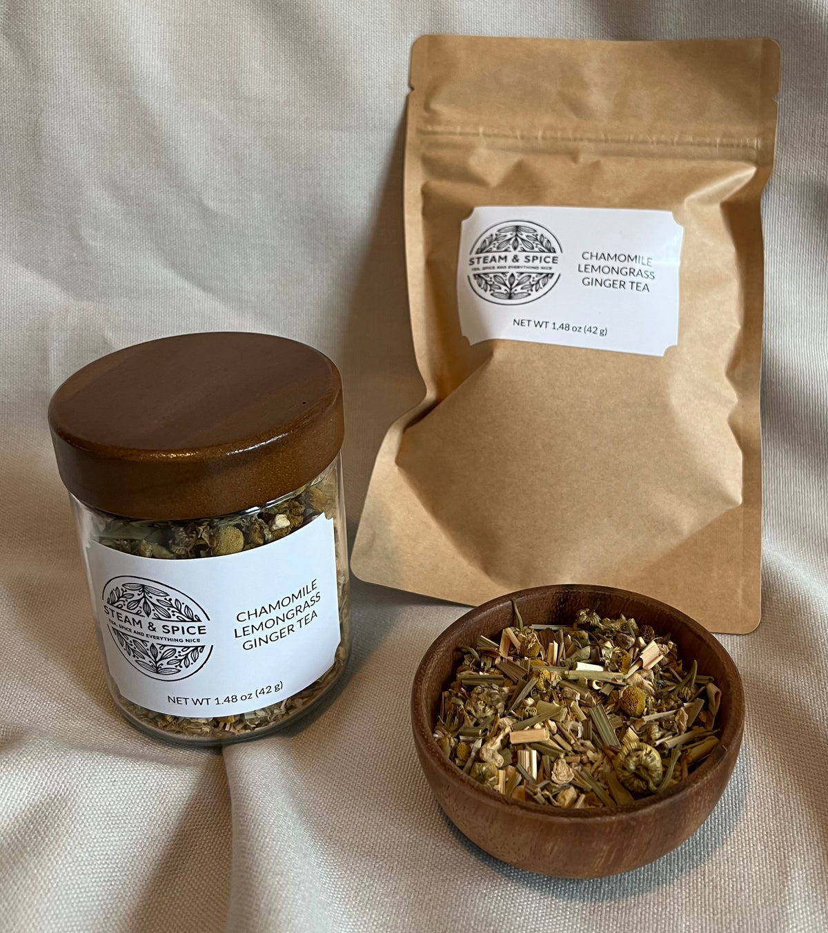 A jar of chamomile lemon ginger tea with a wooden lid, a kraft paper pouch of the same tea, and a small wooden bowl filled with loose tea leaves, arranged on a beige fabric background.