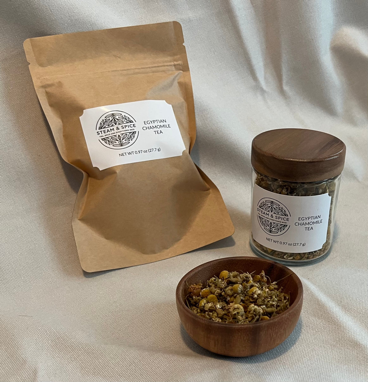 A kraft paper pouch and a glass jar with a wooden lid, both labeled Egyptian Chamomile Tea, arranged on a draped cream fabric background with a small wooden bowl of loose chamomile flowers in front.