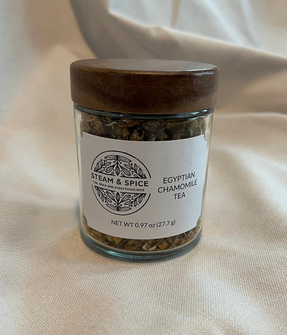 A clear glass jar of Steam & Spice Egyptian Chamomile Tea with a dark brown wooden lid, set against a draped beige fabric background.