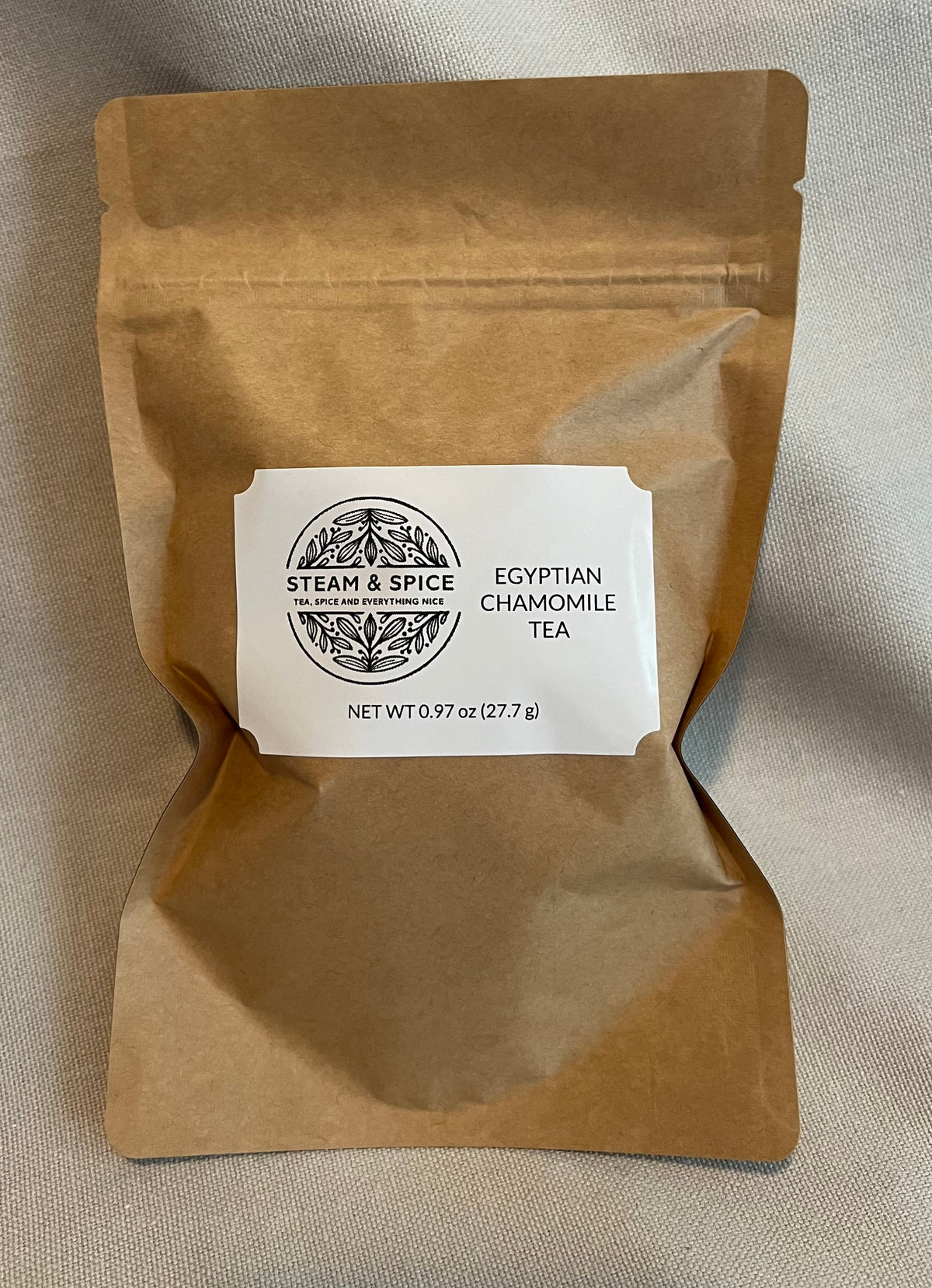 A brown kraft paper stand-up pouch of Steam & Spice Egyptian Chamomile Tea, featuring a white label in the center, set against a neutral beige fabric background.