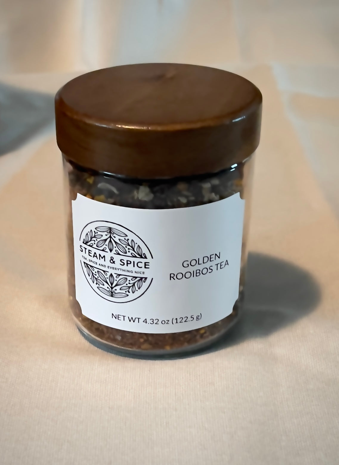 A glass jar of Steam & Spice Golden Rooibos Tea with a dark brown wooden lid and a white paper label, set against a neutral beige fabric background.