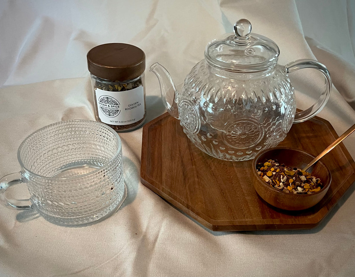 A clear textured glass teapot with a matching glass mug and a small glass jar of loose-leaf herbal tea on a wooden tray, alongside a small wooden bowl of tea blend and a spoon.