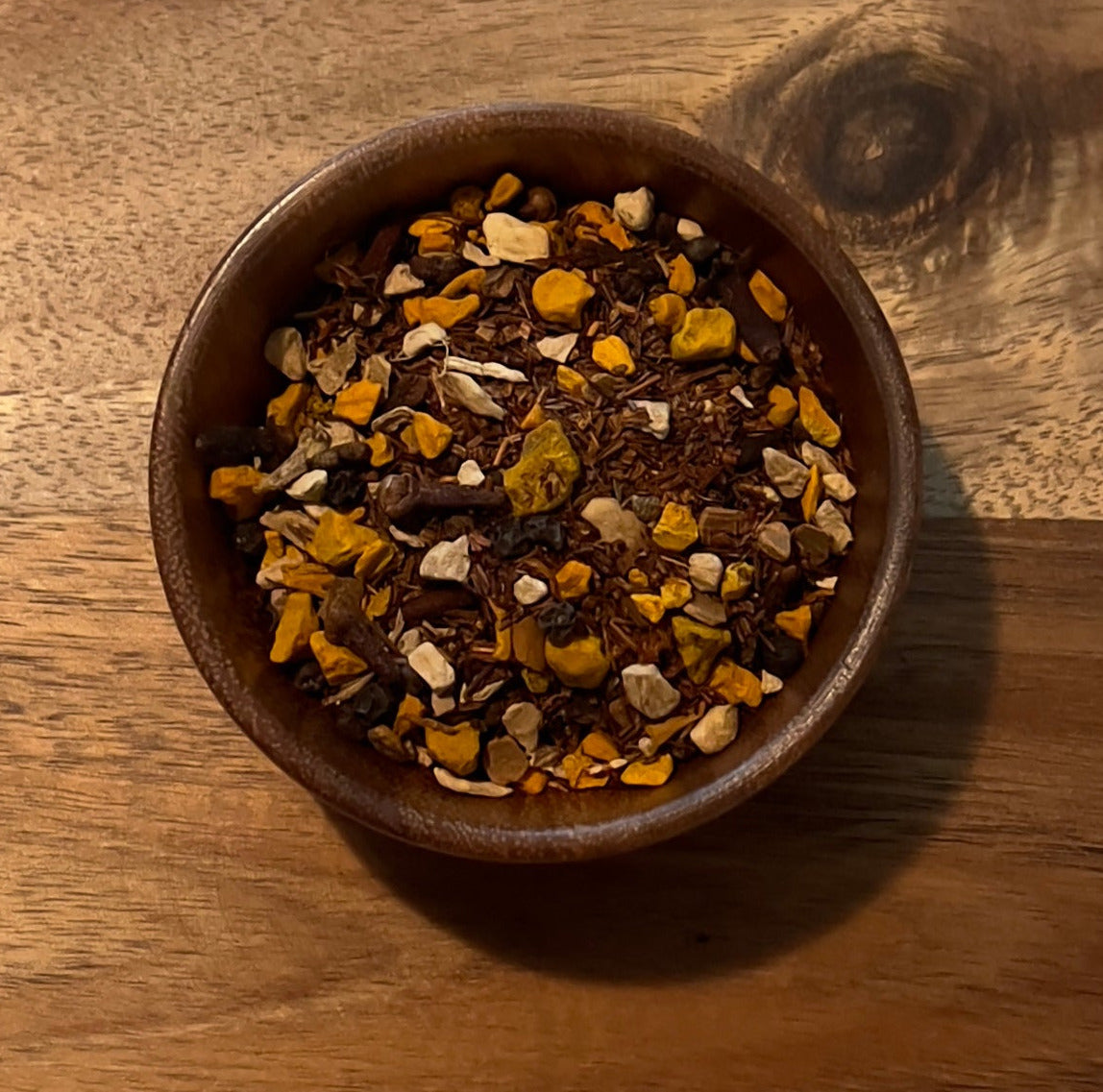 A small brown wooden bowl filled with a colorful mix of dried herbs, seeds, and spices, placed in the center of a large octagonal wooden serving tray on a light fabric background.