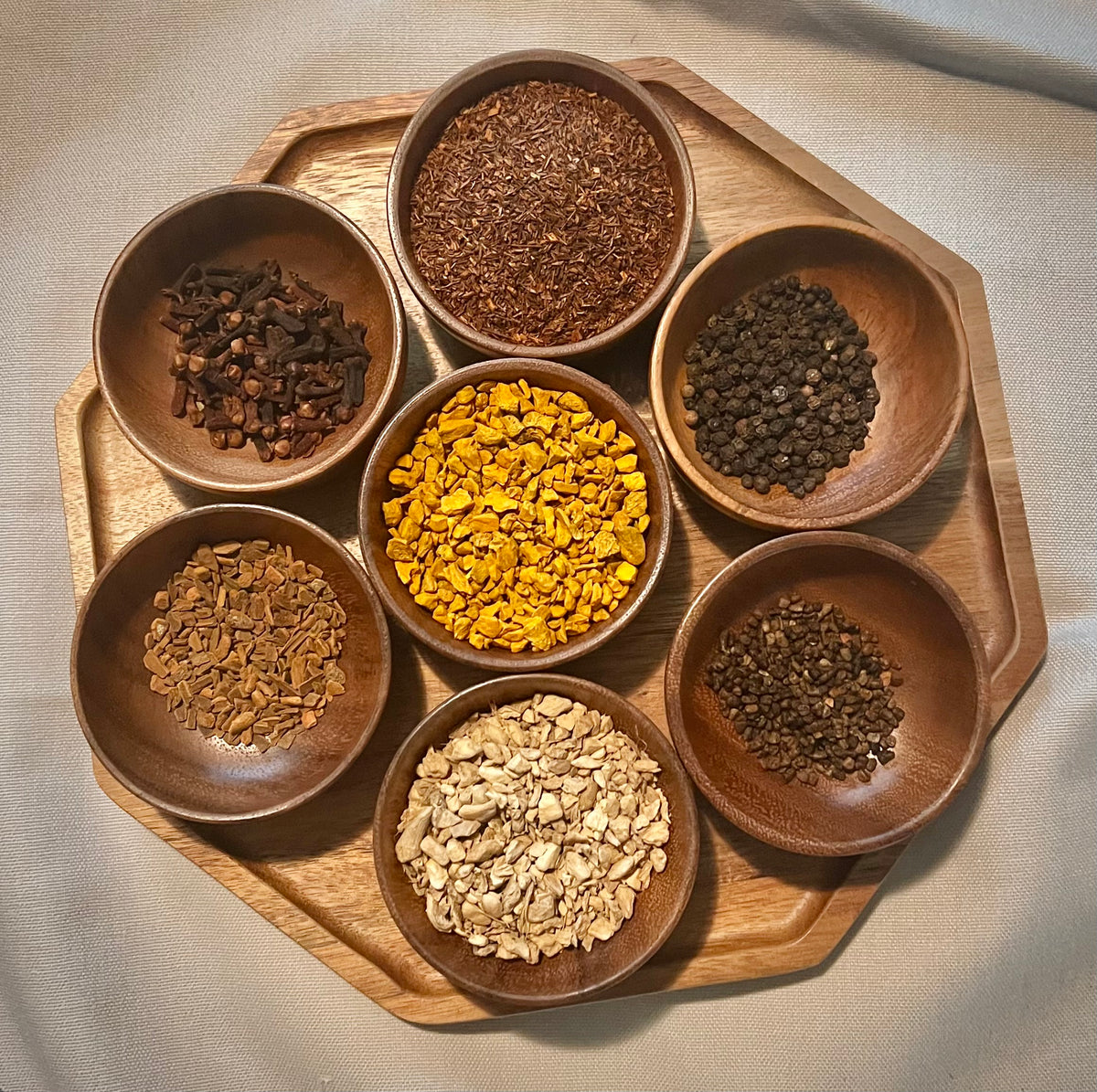 Six small wooden bowls arranged on a larger octagonal wooden tray, each containing different dried botanicals or spices in varied earthy colors, photographed from above on a light fabric surface.