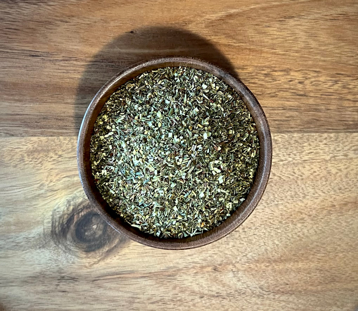 A top-down view of a small brown wooden bowl filled with dried green rooibos leaves, set on a wooden tabletop.