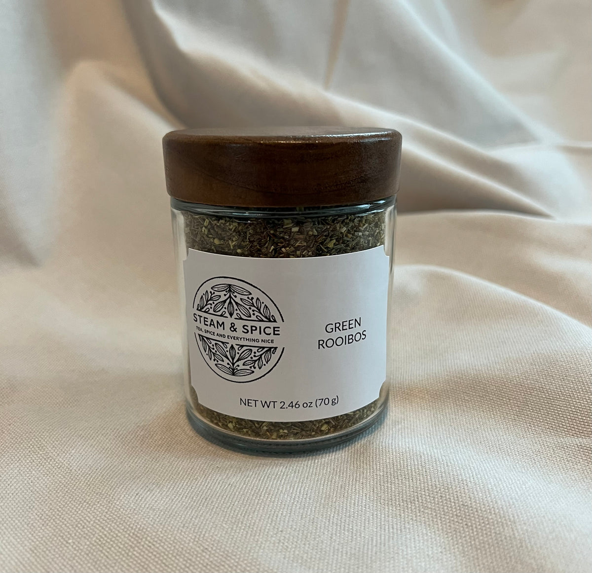 A clear glass jar of Steam & Spice Green Rooibos spice blend with a dark wooden lid, set against a draped cream fabric background.
