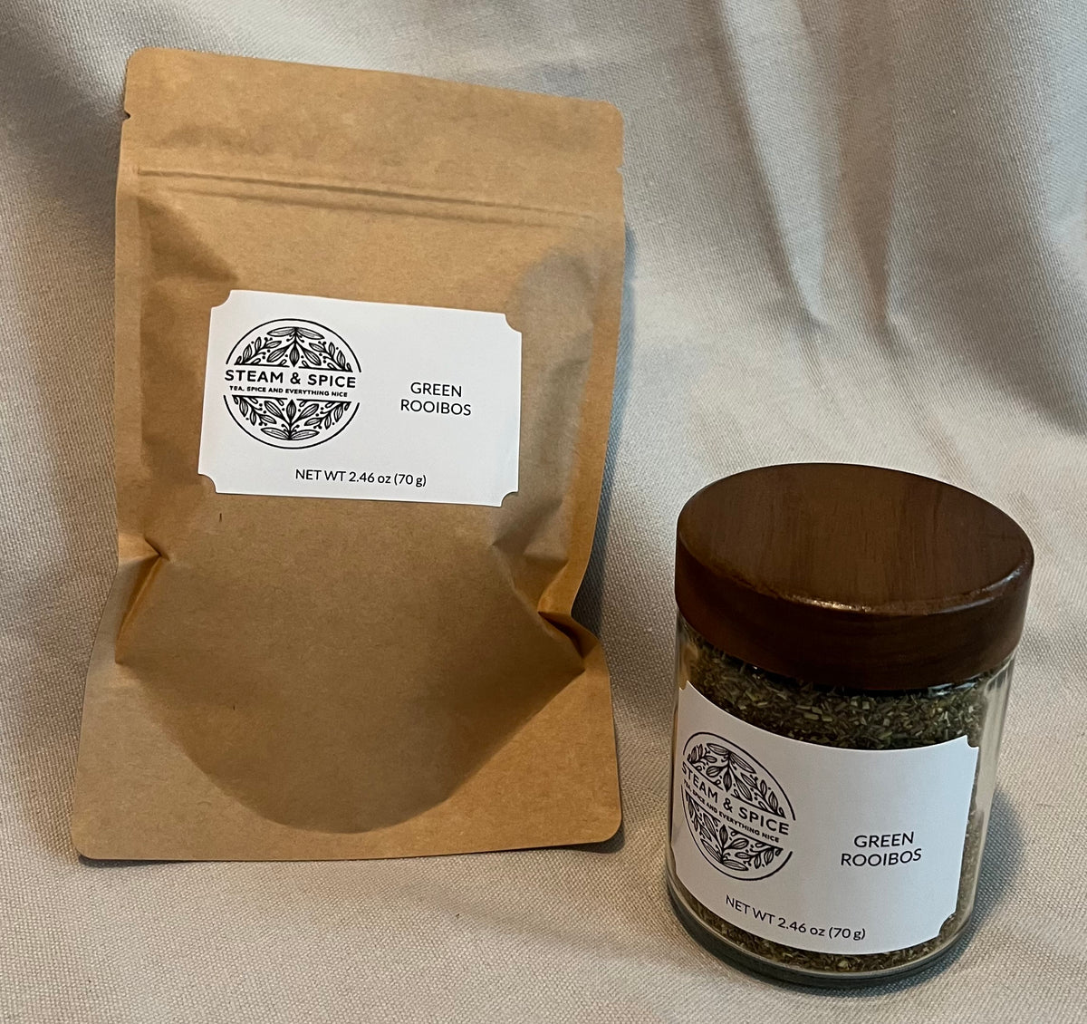 A kraft paper pouch and a small glass jar with a dark wooden lid, both labeled Steam & Spice Green Rooibos, placed on a beige fabric background.