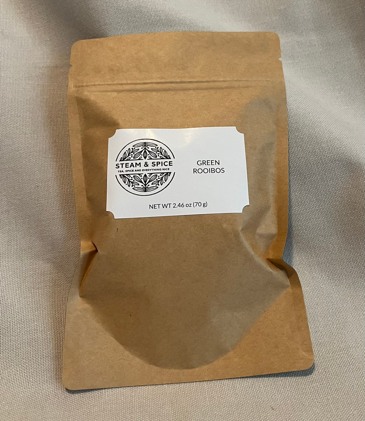 A brown kraft paper stand-up pouch with a white label reading 'Steam & Spice' and 'Green Rooibos', photographed on a light fabric background.