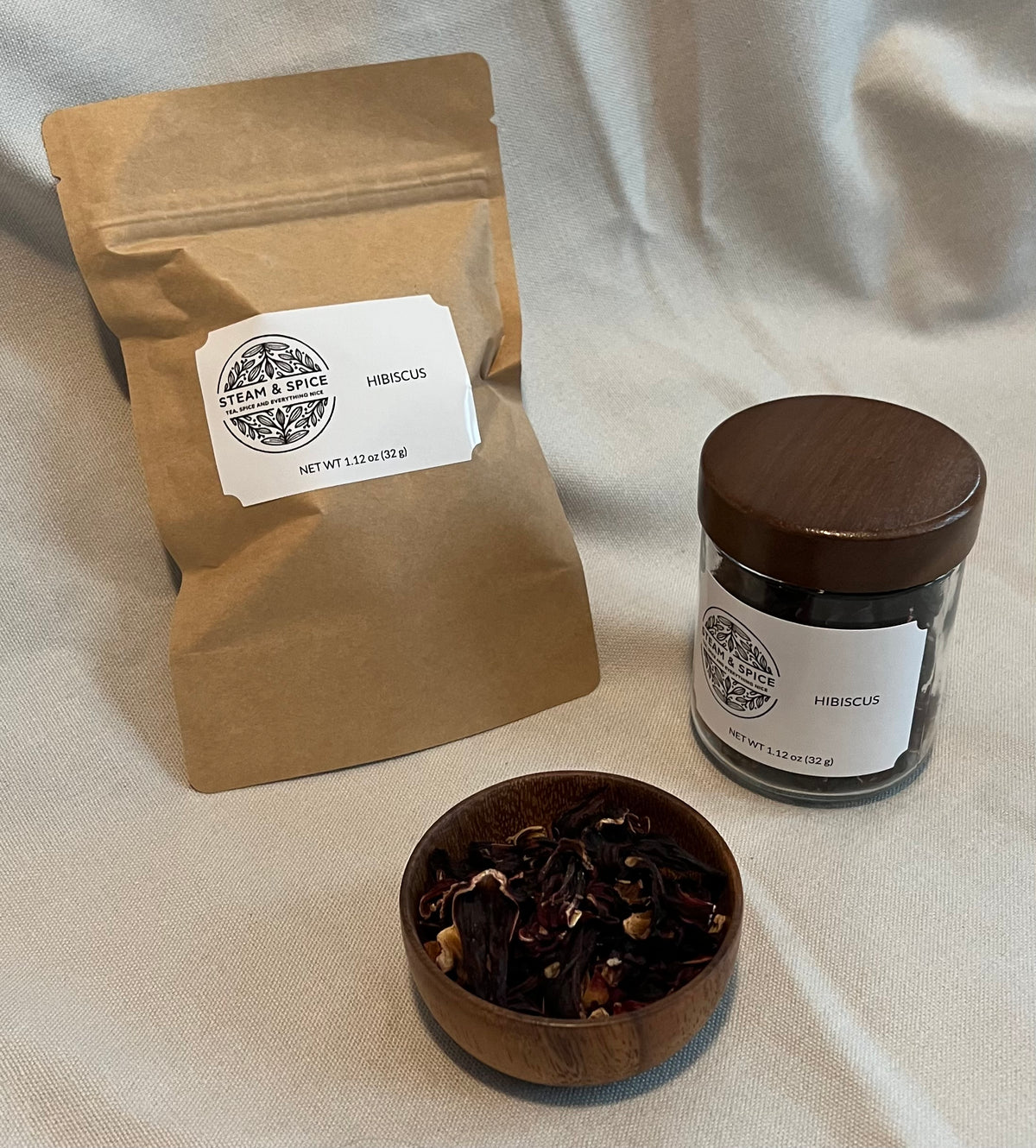 A kraft paper pouch labeled Hibiscus and a small glass jar with a dark wooden lid, both from the brand Tea & Spice, arranged on a draped cream fabric background with a small wooden bowl of loose hibiscus flowers in front.