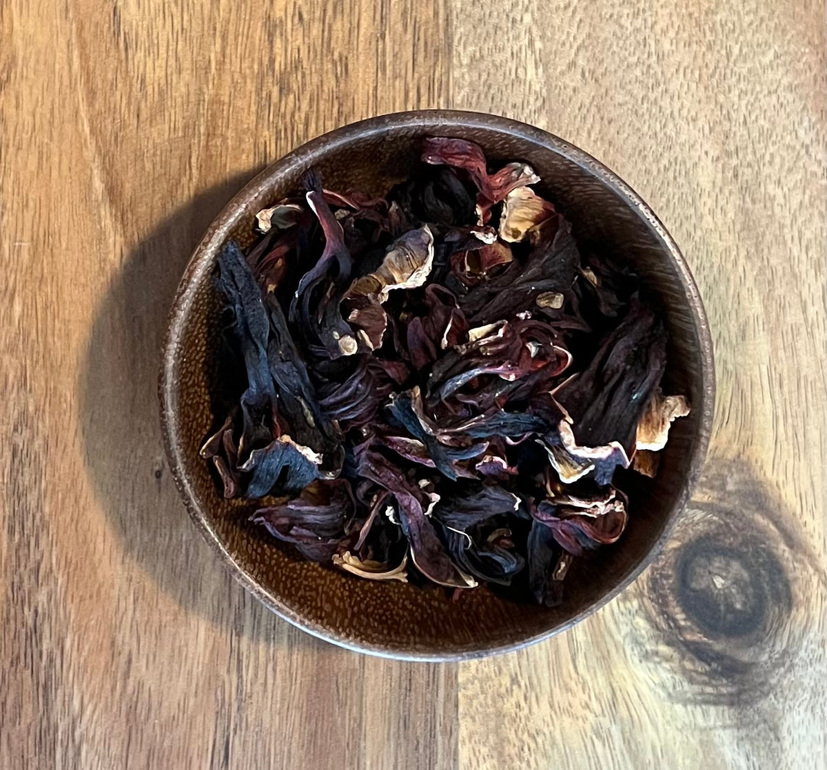 Dried hibiscus flowers in a small brown ceramic bowl, placed on a wooden tabletop.