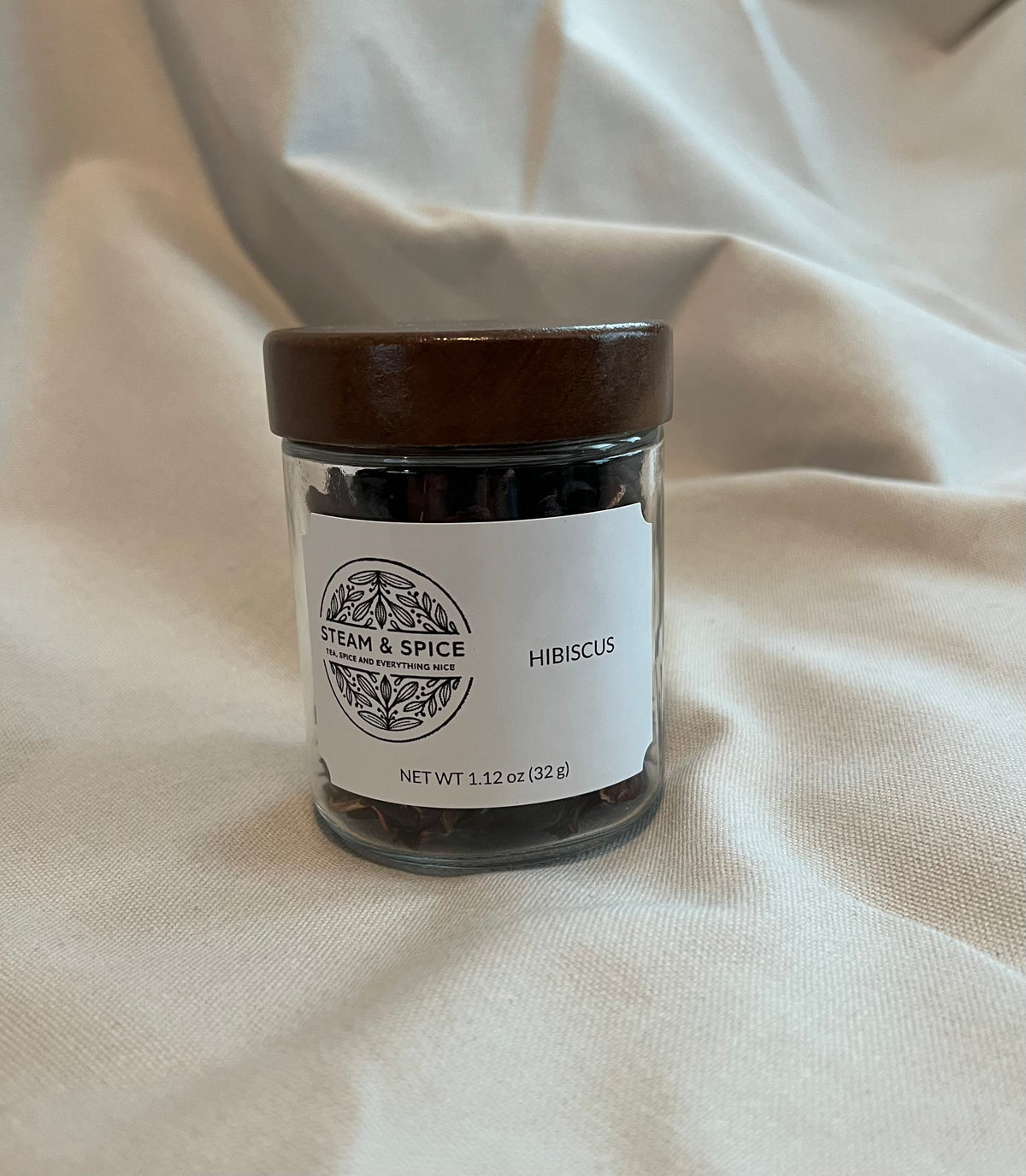 A small clear glass jar filled with dark dried hibiscus flowers, topped with a dark brown wooden lid and labeled 'Steam & Spice' and 'Hibiscus', set against a draped cream fabric background.