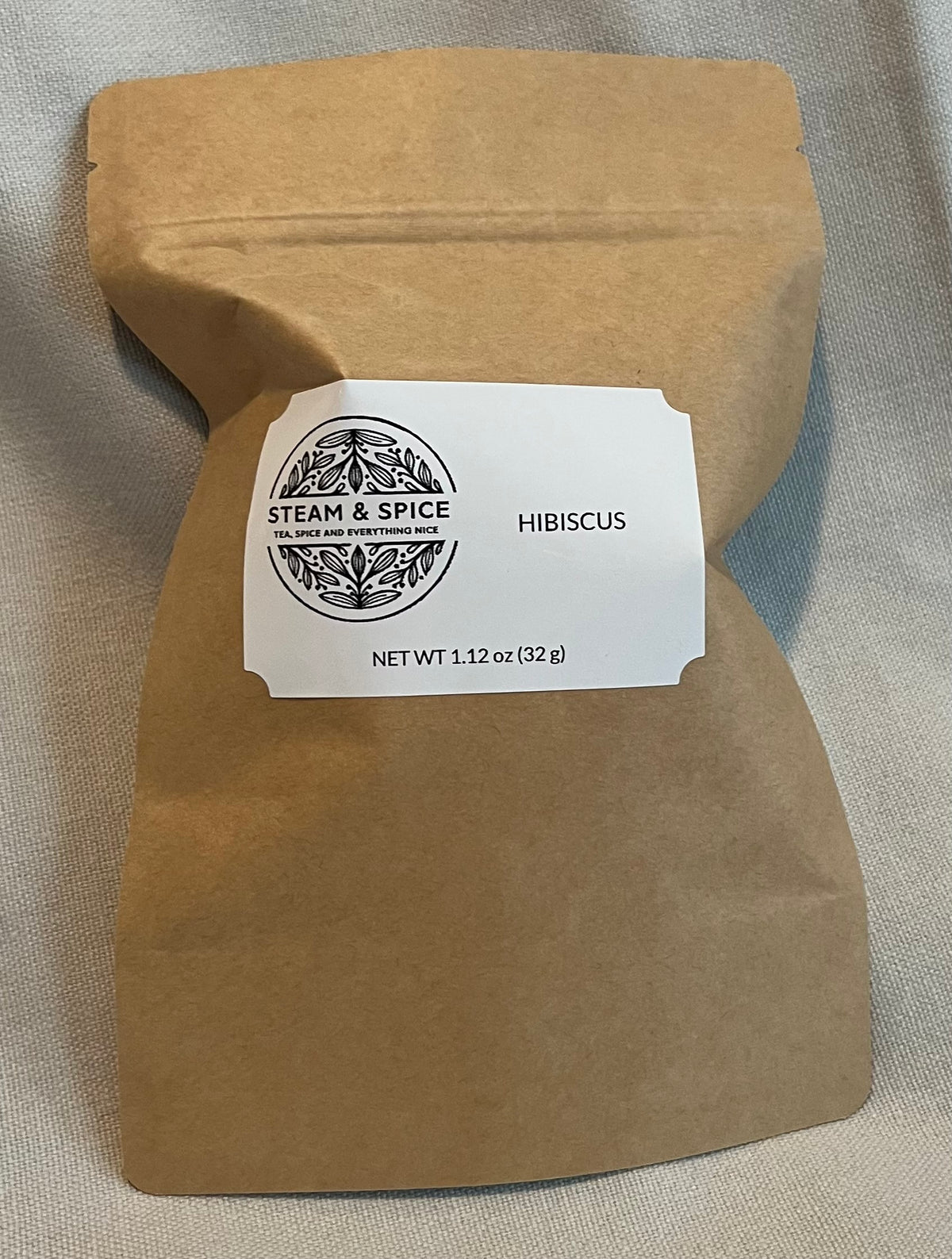 A brown kraft paper stand-up pouch with a white label reading 'STEAM & SPICE' and 'HIBISCUS', photographed on a light fabric background.