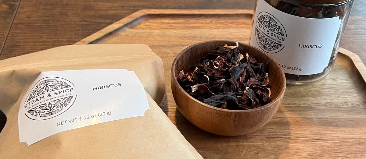 A kraft paper pouch labeled 'Steam & Spice Hibiscus' and a clear jar labeled 'HIBISCUS' sit on a wooden tray beside a small wooden bowl filled with dried hibiscus flowers.
