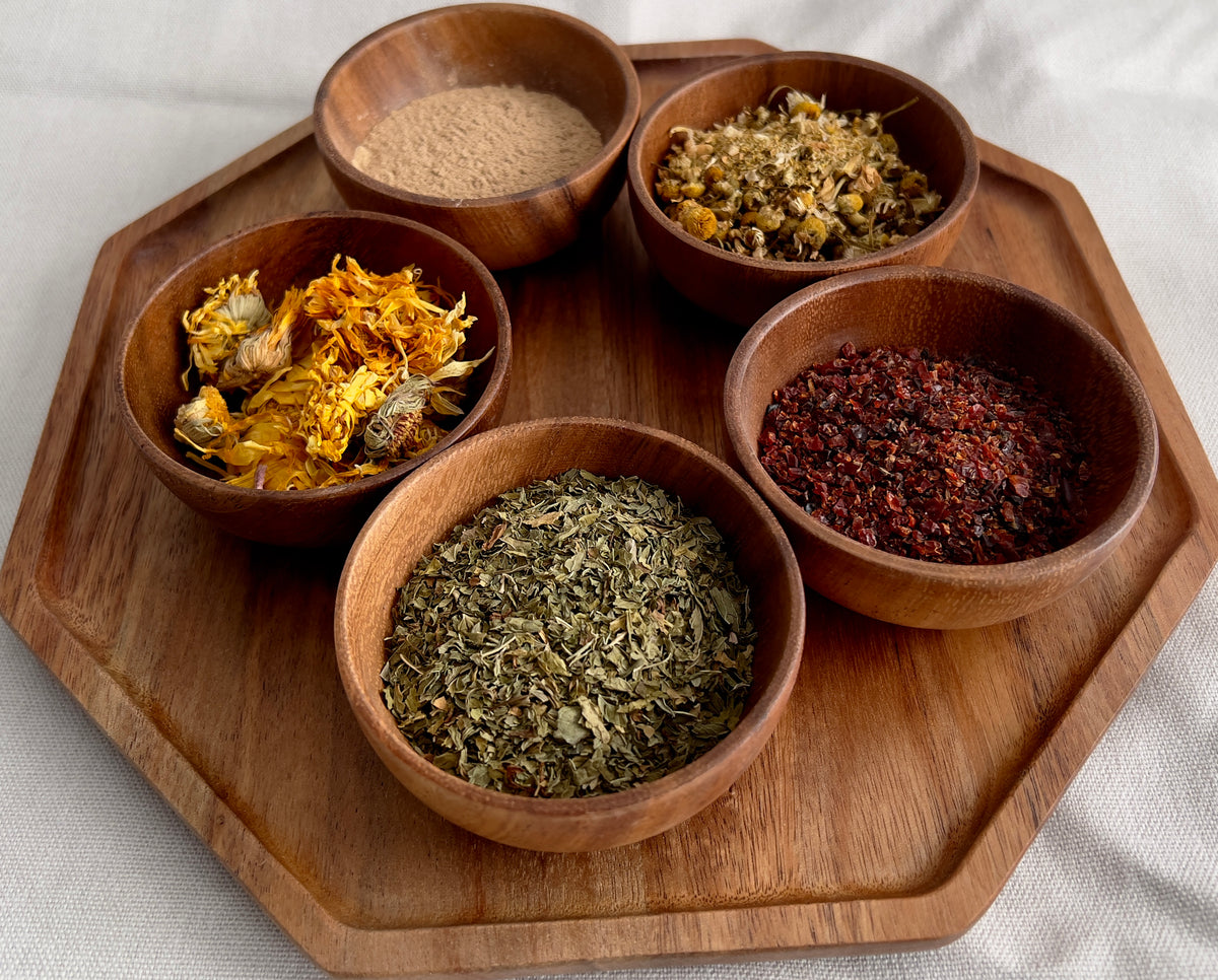 Five wooden bowls with different dried herbs and spices on a wooden tray.