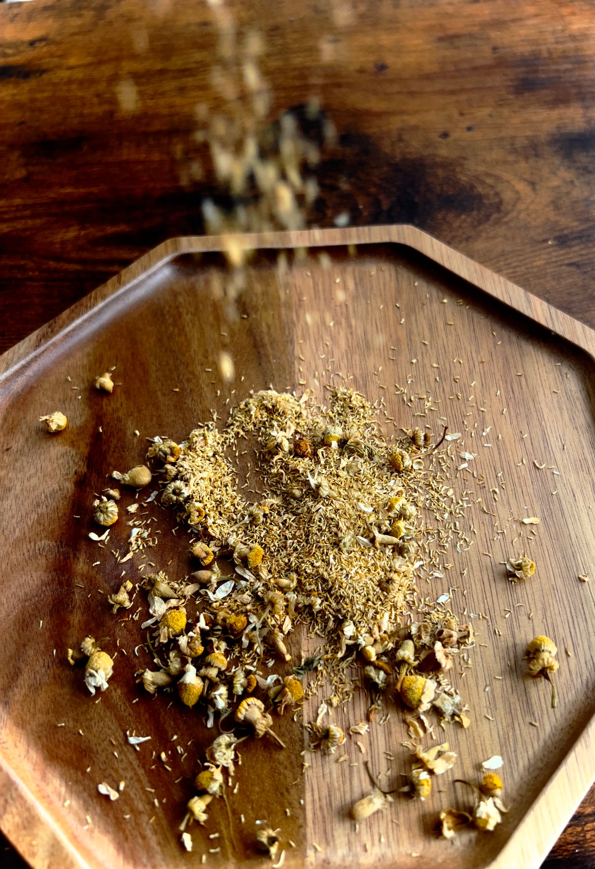 Wooden bowl with dried chamomile herbs being sprinkled onto a wooden surface