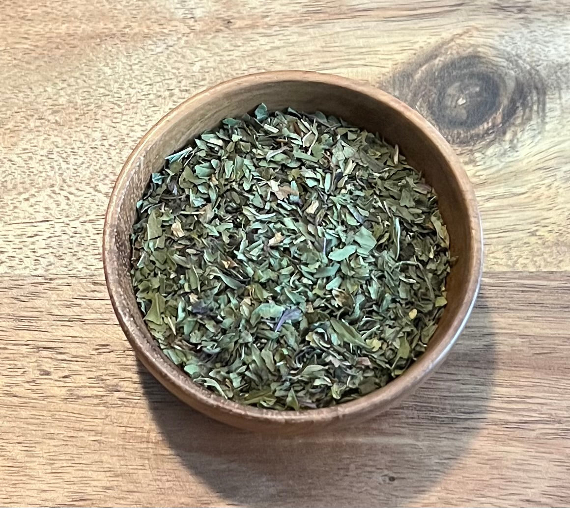 Dried green peppermint leaves in a small round wooden bowl, placed on a wooden tabletop.