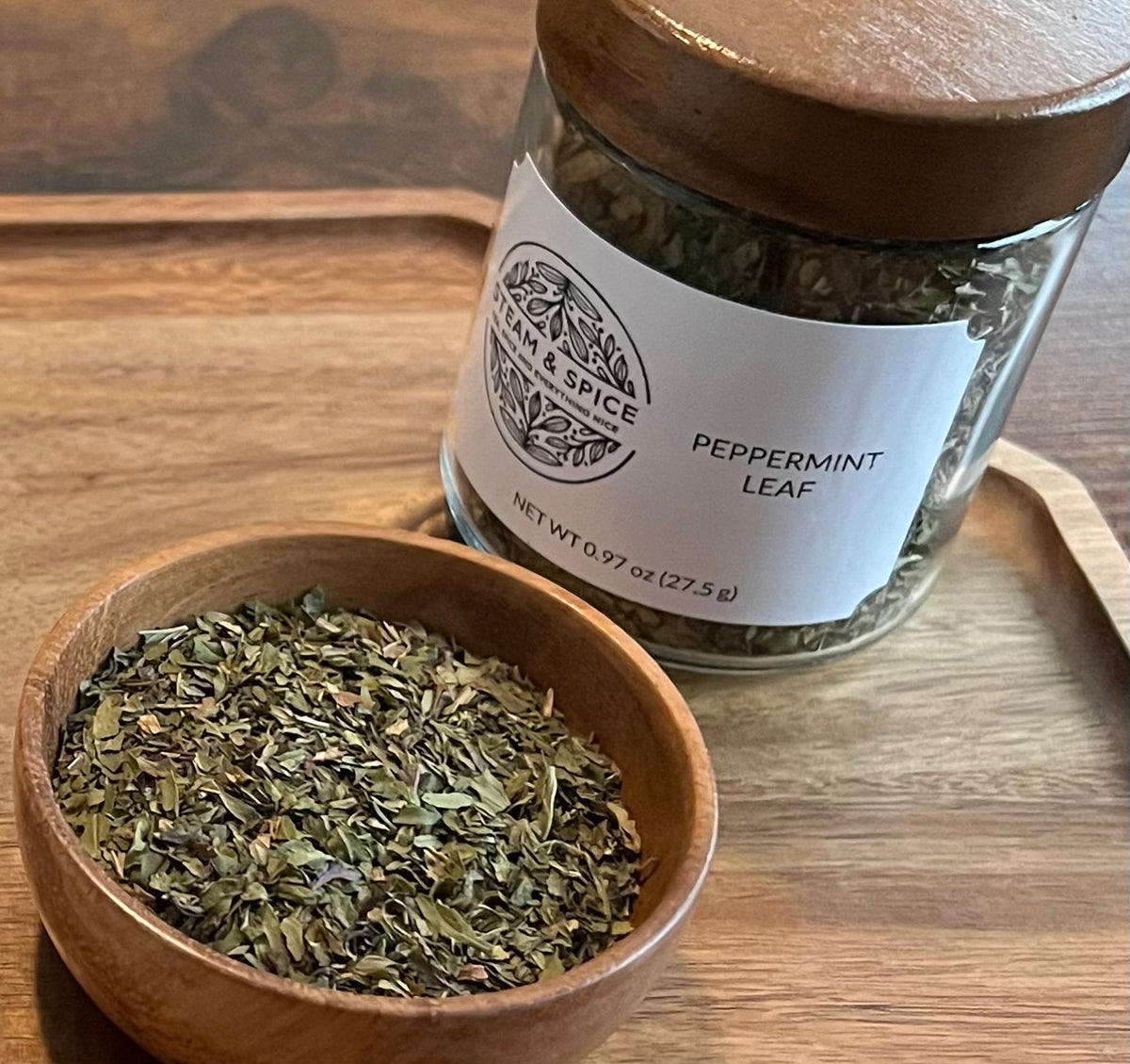 A glass jar labeled 'Peppermint Leaf' with a wooden lid sits on a wooden surface next to a small wooden bowl filled with loose dried peppermint leaves.