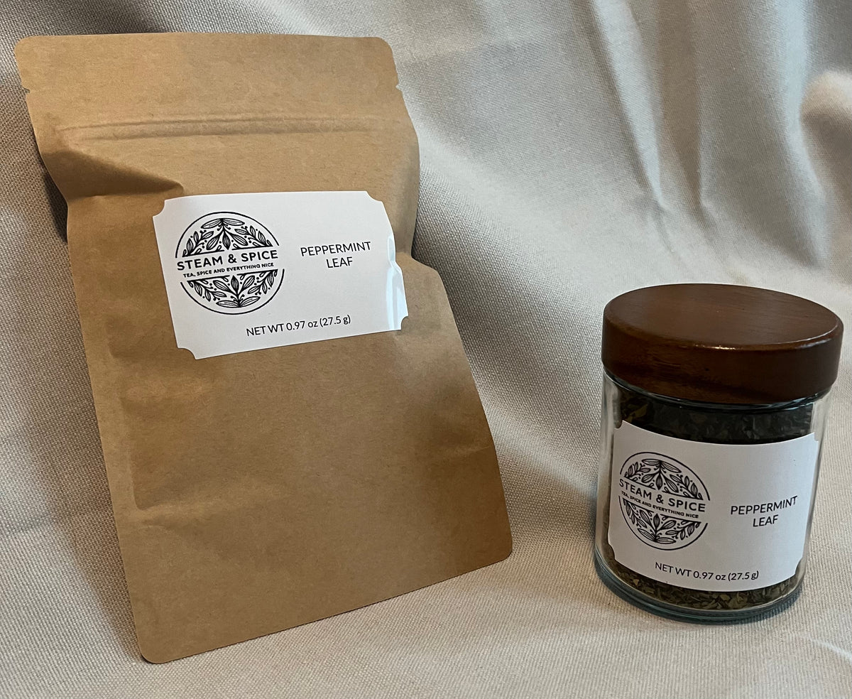 A kraft paper pouch and a small glass jar of peppermint leaf from Steam & Spice, displayed on a draped cream fabric background.