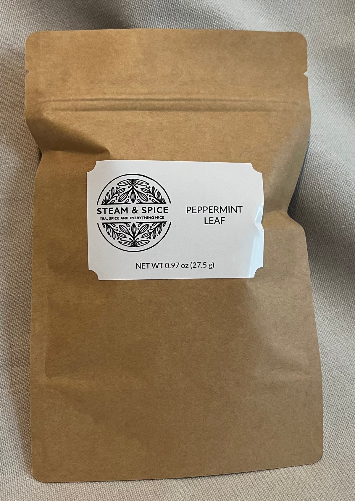 A brown kraft paper stand-up pouch with a white label reading 'Steam & Spice' and 'Peppermint Leaf' on a light fabric background.