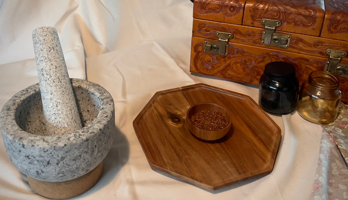 A stone mortar and pestle set on a white fabric surface, alongside a wooden octagonal tray, a small wooden bowl of spices, two glass jars, and an ornate carved wooden chest.