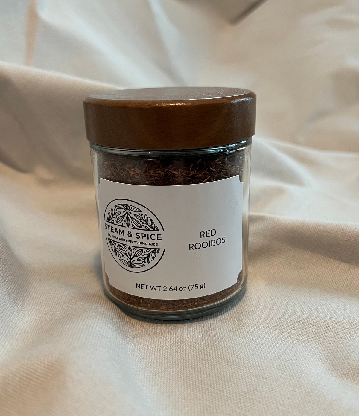 A glass jar of Steam & Spice Red Rooibos tea with a brown wooden lid and a white label, set against a draped beige fabric background.