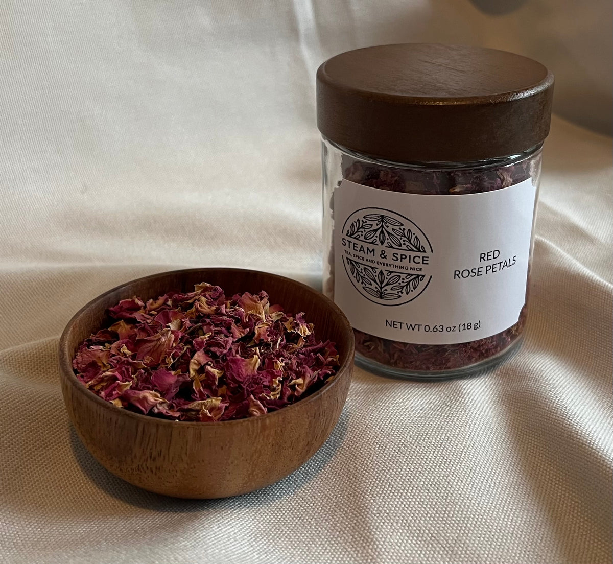 A glass jar of red rose petals with a dark wooden lid and a white label reading 'STEAM & SPICE' and 'RED ROSE PETALS', shown beside a small wooden bowl filled with dried red rose petals on a cream fabric background.
