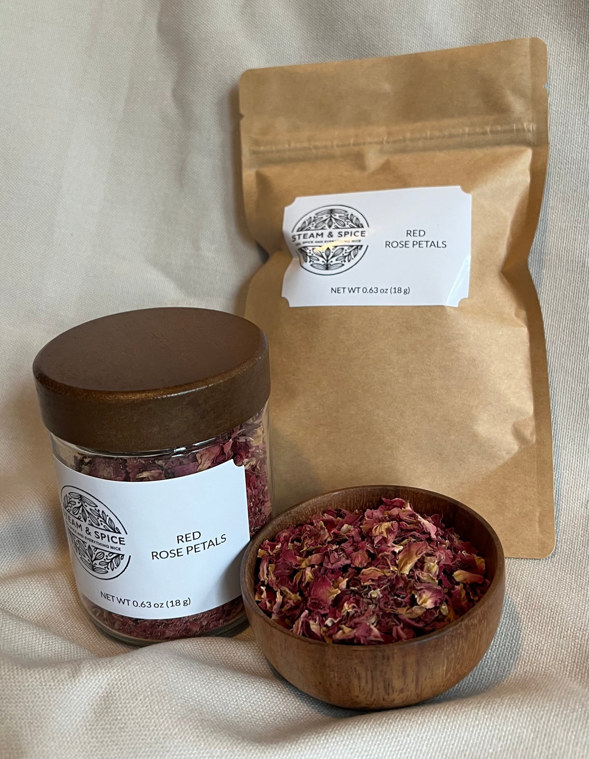 A jar of dried red rose petals with a wooden lid, a kraft paper pouch, and a small wooden bowl filled with loose dried rose petals, arranged on a beige fabric background.