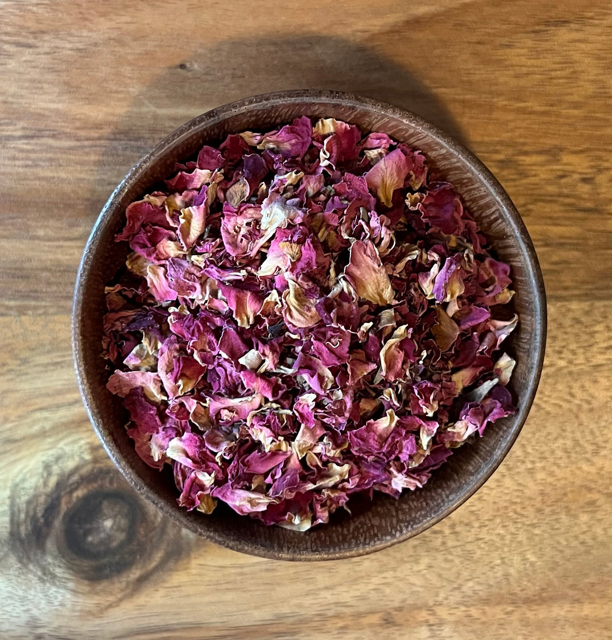Dried rose petals in a dark brown wooden bowl, photographed from above on a wooden tabletop.