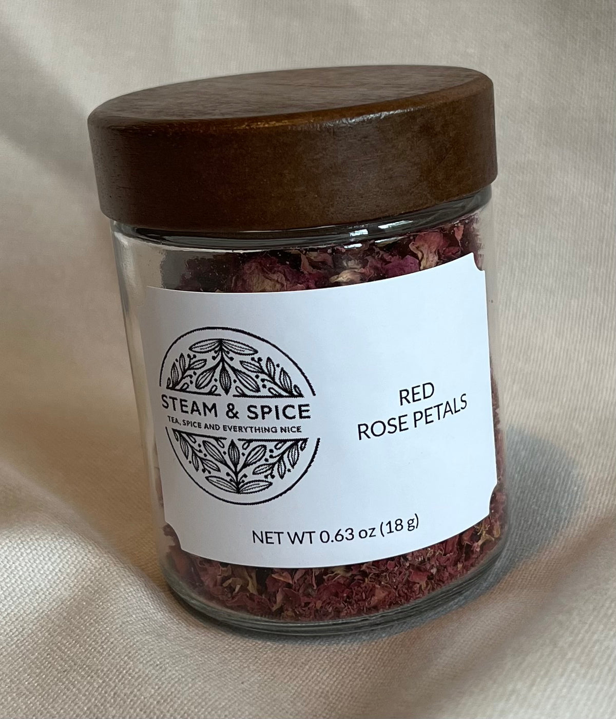 A clear glass jar filled with dried red rose petals, featuring a dark brown wooden lid and a white paper label that reads 'STEAM & SPICE' and 'RED ROSE PETALS'. The jar is set against a draped beige fabric background.