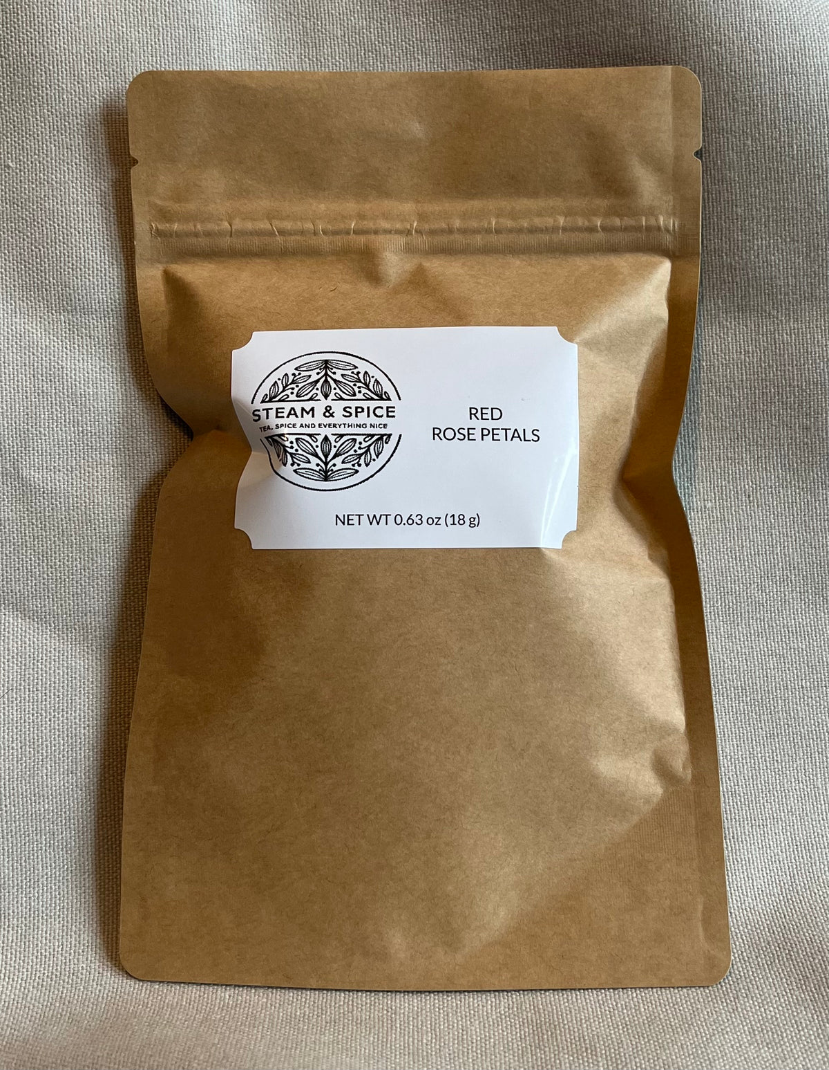 A brown kraft paper stand-up pouch with a white label reading 'STEAM & SPICE' and 'RED ROSE PETALS', photographed on a light beige fabric background.
