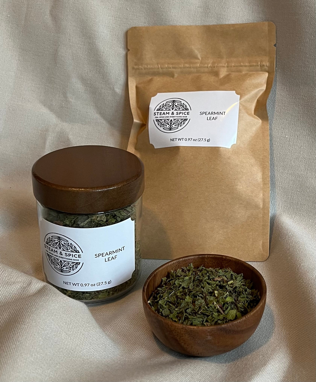 A jar of spearmint leaf tea with a wooden lid, a kraft paper pouch, and a small wooden bowl filled with loose spearmint leaves, arranged on a beige fabric background.