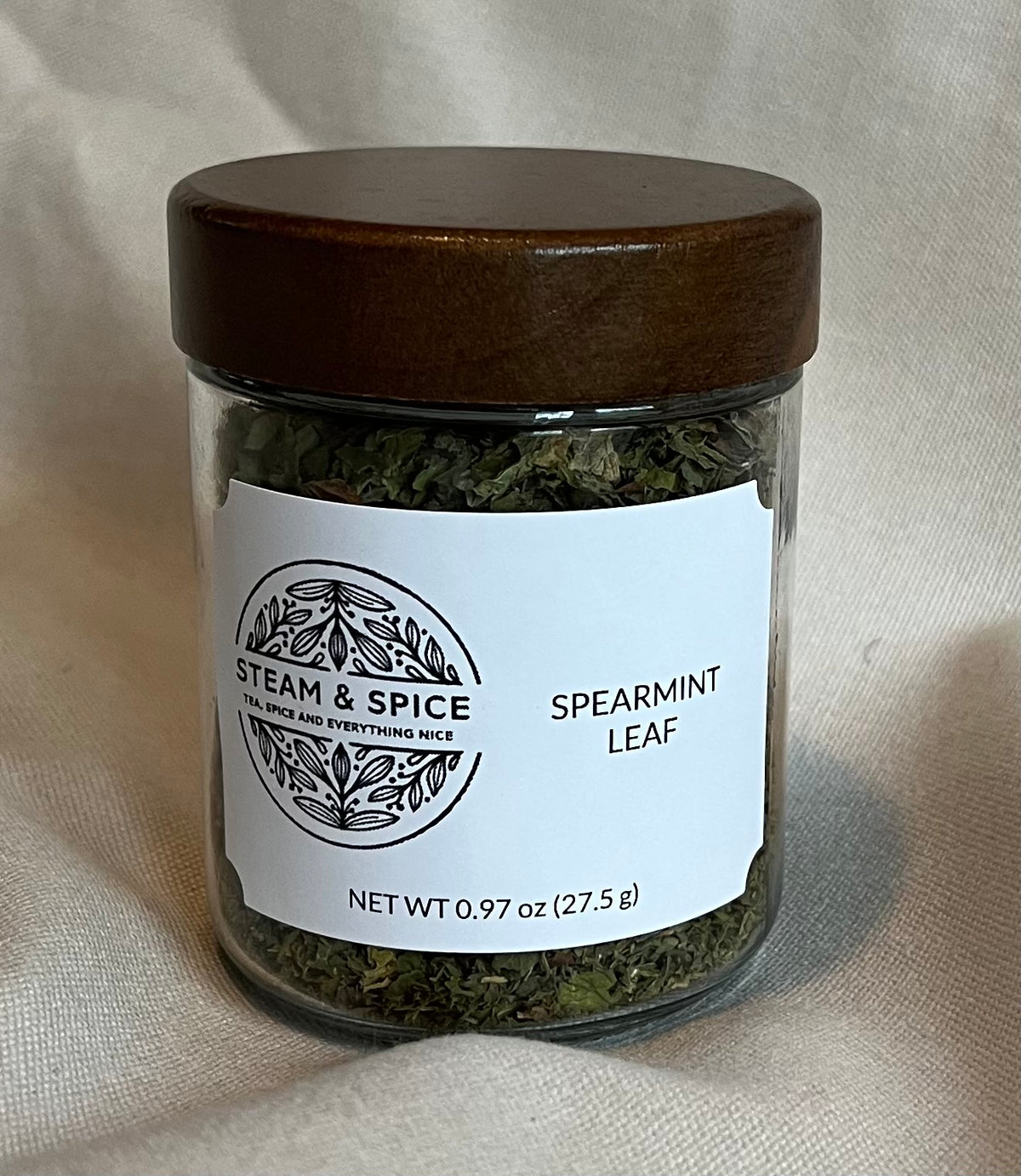 A clear glass jar filled with dried spearmint leaf tea, featuring a dark brown wooden lid and a white label that reads 'STEAM & SPICE' and 'SPEARMINT LEAF'. The jar is set against a neutral beige fabric background.