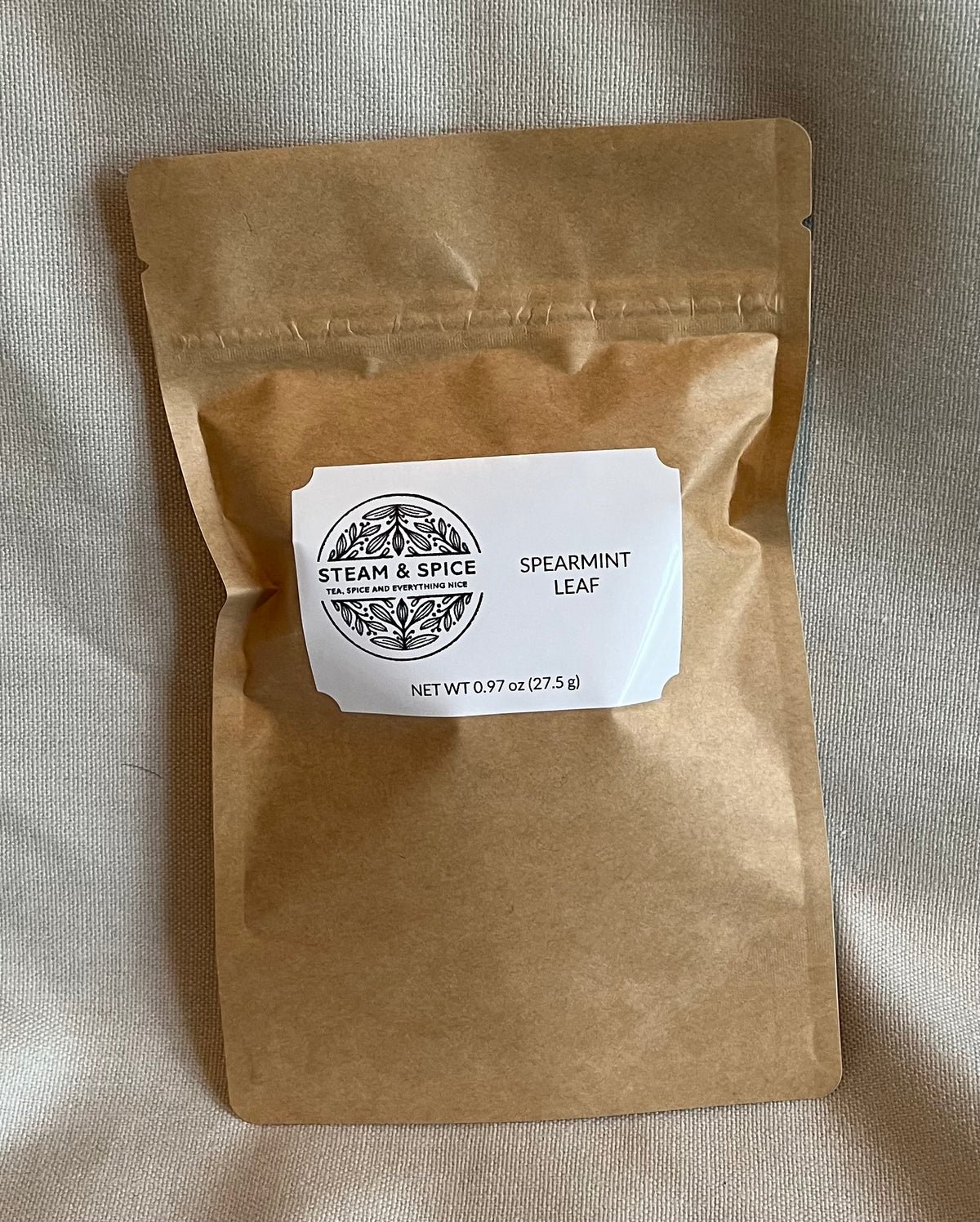 A brown kraft paper stand-up pouch with a white label reading 'STEAM & SPICE' and 'SPEARMINT LEAF', placed on a beige fabric background.